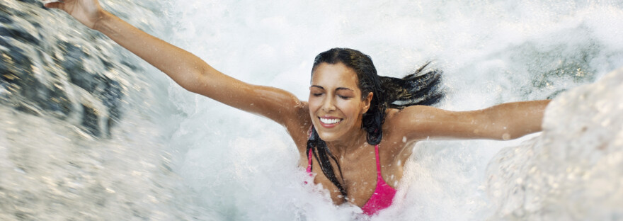 Eine Frau steht lachend unter einem Wasserfall.. Voller Glück breitet sie die Arme aus.
